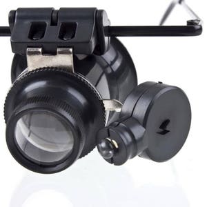 K&ouml;nnte beinhalten: Eine schwarze Lupe mit eingebautem LED-Licht, befestigt an einem Kopfband. Die Lupe hat eine gro&szlig;e Linse und eine kleinere Linse an der Seite. Das Kopfband ist verstellbar und hat einen schwarzen Kunststoffrahmen.