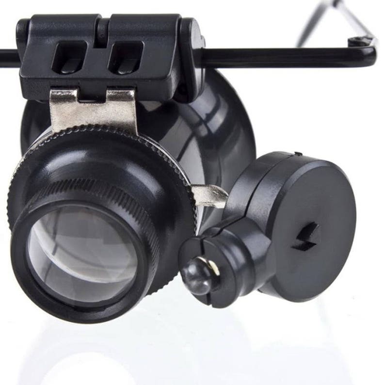 K&ouml;nnte beinhalten: Eine schwarze Lupe mit eingebautem LED-Licht, befestigt an einem Kopfband. Die Lupe hat eine gro&szlig;e Linse und eine kleinere Linse an der Seite. Das Kopfband ist verstellbar und hat einen schwarzen Kunststoffrahmen.