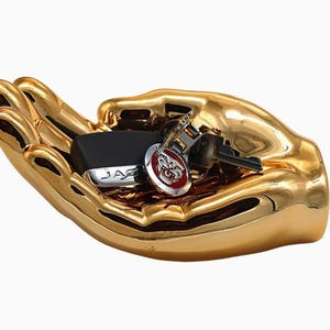 May include: A gold-colored, hand-shaped decorative tray holding a black car key fob with the word "JAG" and a set of keys with a red emblem. The tray has a glossy finish and is set against a white background.