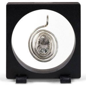 Pode incluir: Uma gema oval de vidro transparente está envolta em uma espiral prateada, exibida em uma moldura quadrada preta com uma janela circular transparente.