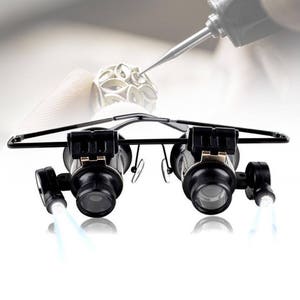 K&ouml;nnte beinhalten: Schwarze Lupenbrille mit eingebauten LED-Lichtern, die f&uuml;r den freih&auml;ndigen Gebrauch konzipiert ist. Die Brille hat ein Kopfband und verstellbare Linsen.