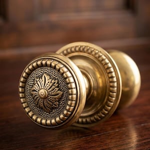 May include: A close-up shot of a brass door knob featuring an intricate floral pattern. The knob is circular with a detailed edge design. The background is a dark wooden surface.
