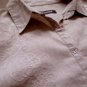 May include: A light beige linen shirt with embroidered floral details. The shirt has a button-up front and a collar with the text "hawkhead" on the inside.