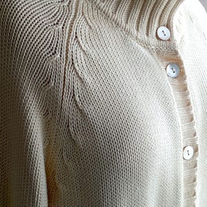 May include: Cream-coloured knitted cardigan with a high, ribbed collar and button closure. The sweater features a detailed knit pattern on the shoulders and sleeves, and is made of a soft, textured material.