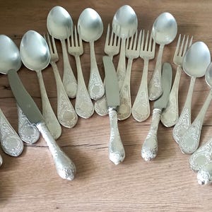May include: An assortment of antique silverware, including spoons, forks, and knives, arranged on a wooden surface. The silverware has ornate, detailed handles and a shiny, silver finish. The collection includes various sizes of spoons, forks, and knives.