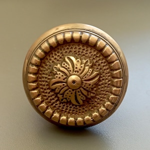 May include: An antique brass doorknob with a floral centre. The round knob has a textured surface and a decorative border. The colour is a warm, aged brass.