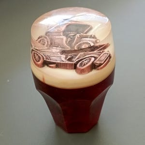 May include: A gear shift knob featuring a clear resin top section that encapsulates a metallic copper-coloured vintage car model. The base of the knob is made of a solid, translucent dark red material with a faceted, multi-sided geometric shape.