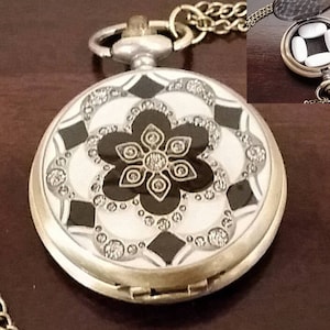 May include: A silver pocket watch with a black and white patterned design. The watch has a chain attached to it and opens to reveal a compartment for holding small items.