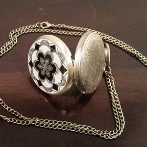 May include: A silver pocket watch with a black and white floral design on the front and an engraved design on the back. The watch is attached to a gold chain.
