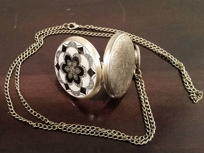 May include: A silver pocket watch with a black and white floral design on the front and an engraved design on the back. The watch is attached to a gold chain.