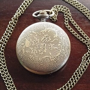 May include: A gold pocket watch with a chain. The watch face has an intricate floral design.