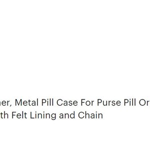 May include: A silver metal pill container with a black and white floral design. The container has a chain attached to it and is on a brown surface.
