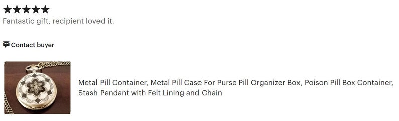 May include: A silver metal pill container with a black and white floral design. The container has a chain attached to it and is on a brown surface.