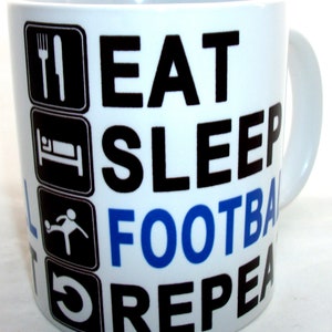 May include: White ceramic mug printed with the words "EAT SLEEP FOOTBALL REPEAT" in black and blue. The mug has black icons of a fork and knife, a bed, a person kicking a ball, and a circular arrow.
