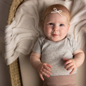 May include: A baby wearing a grey bodysuit and a brown headband with a white bow. The baby is lying in a wicker basket.