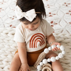 May include: A baby wearing a white bodysuit with a brown rainbow graphic and brown corduroy shorts. The baby is holding a white wooden bead necklace with a brown tassel.