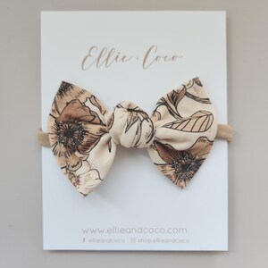 May include: A beige headband with a floral print bow. The bow is tied in a knot and has a brown and beige floral pattern. The headband is on a white card with the text "Ellie Coco" and "www.ellieandcoco.com".