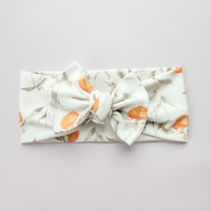 May include: A white headband with a knotted bow, featuring a floral print of orange blossoms and leaves.