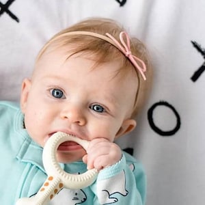 May include: A baby wearing a light blue onesie with a white polar bear design is chewing on a white and brown teething ring. The baby is wearing a light brown headband with a pink bow.