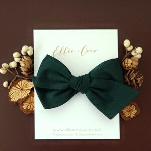 May include: A dark green fabric bow with a simple design. The bow is on a white card with the text "Ellie Coco" and the website address "www.ellieandcoco.com".