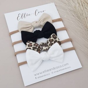 May include: Set of three hair bows with nude, black, and leopard print fabric. The bows are tied with a knot in the centre and are attached to nude elastic headbands. The card reads "Ellie + Coco" and includes the website address "www.ellieandcoco.com".