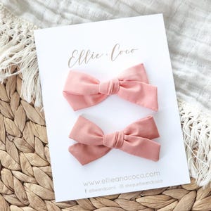 May include: Two pink fabric hair bows on a white card with the text "Ellie + Coco" and website information. The bows are displayed on a woven wicker surface with a white fabric background.