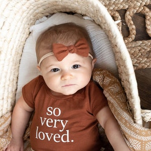May include: A baby wearing a brown bodysuit with the text "so very loved." The baby is wearing a brown bow headband and is lying in a woven basket.