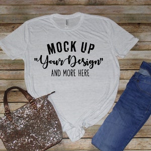 May include: A light gray heathered short-sleeve t-shirt with black text that reads MOCK UP Your Design AND MORE HERE, paired with a pair of blue denim jeans and a small handbag covered in shimmering bronze-colored sequins and metallic handles.