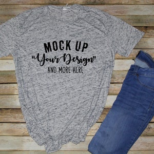 May include: A heather gray short-sleeve t-shirt with black text that reads MOCK UP "Your Design" AND MORE HERE, paired with a pair of blue denim jeans. The shirt features a speckled fabric texture and a crew neckline, while the jeans are dark blue.