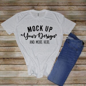 May include: A light gray heathered short-sleeve t-shirt featuring the black printed text MOCK UP "Your Design" AND MORE HERE, paired with a pair of blue denim jeans. The t-shirt has a crew neckline and a standard fit, while the jeans are folded.