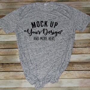 May include: A heather gray short-sleeve t-shirt with black text printed on the front that reads MOCK UP "Your Design" AND MORE HERE. The fabric has a speckled, marled appearance with a crew neckline and a standard fit.
