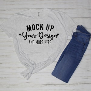 May include: A light gray heathered short-sleeve t-shirt with a knotted hem and black text that reads MOCK UP "Your Design" AND MORE HERE, paired with a pair of dark blue denim jeans. The shirt features a crew neckline and a soft, speckled fabric.