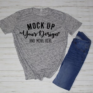 May include: A heather gray short-sleeve t-shirt with black text that reads MOCK UP "Your Design" AND MORE HERE, paired with a pair of blue denim jeans. The t-shirt features a speckled fabric pattern and a crew neckline, displayed flat.