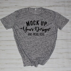May include: A heather gray short-sleeve t-shirt with black text printed on the front that reads MOCK UP "Your Design" AND MORE HERE. The fabric has a mottled, speckled appearance with a crew neckline and standard hemmed sleeves and bottom.