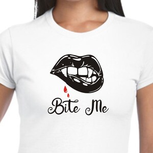 May include: White t-shirt with a black graphic of vampire lips, fangs, and a drop of red blood. The words "Bite Me" are written in a cursive font below the graphic. The t-shirt has a crew neck.