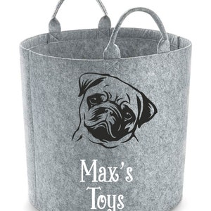 Può includere: Un cesto portaoggetti in feltro grigio con due manici. Sul davanti è presente una grafica nera del volto di un carlino, con le parole "Max's Toys" in corsivo bianco sottostante.