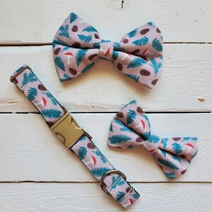 May include: A dog collar and two bow ties featuring a repeating pattern of teal ferns, brown pine cones, and red mushrooms on a light purple background. The collar has a gold-coloured metal buckle and ring. The bow ties are pre-tied.