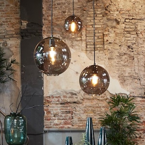 May include: Three pendant lights with spherical, smoky glass shades hang from black cords. The bulbs inside are lit, casting a warm glow. The background features a weathered brick wall, adding an industrial aesthetic to the lighting fixtures.