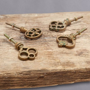 May include: Four antique brass key-shaped cabinet pulls with decorative designs. Each pull has a threaded rod and nut for mounting. The pulls are arranged on a weathered wooden surface, showcasing their vintage aesthetic.