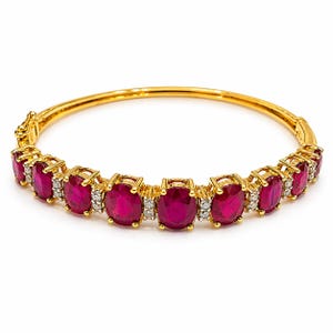 Op de afbeelding: Een gouden armband met ovale, dieprode edelstenen. De armband is geaccentueerd met kleine, heldere stenen en heeft een veilige sluiting. De edelstenen zijn gezet in individuele gouden zettingen.