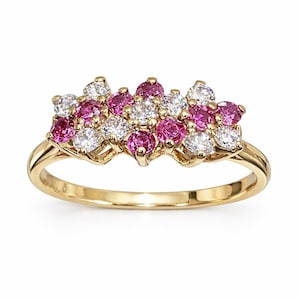 Può includere: Un anello d'oro con un gruppo di pietre preziose rosa e bianche. Le pietre preziose sono disposte a motivo floreale sulla parte superiore dell'anello. La fascia è in oro lucido semplice.