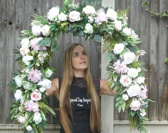 May include: A woman is holding up a large floral arch made of white and pink flowers and greenery. The arch is in the shape of a half-circle and is decorated with roses, hydrangeas, and other flowers. The woman is wearing a black apron with the text "Special Day Bouquets Kent" on it.