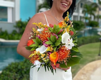 May include: A bride in a white wedding dress smiles while holding a bouquet of orange, yellow, and white flowers. The bouquet includes calla lilies, ginger, and other tropical flowers.