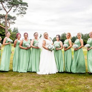 May include: A group of bridesmaids in sage green dresses stand in a line on a golf course. The bride is in the center wearing a white wedding dress. The bridesmaids are holding bouquets of white flowers.