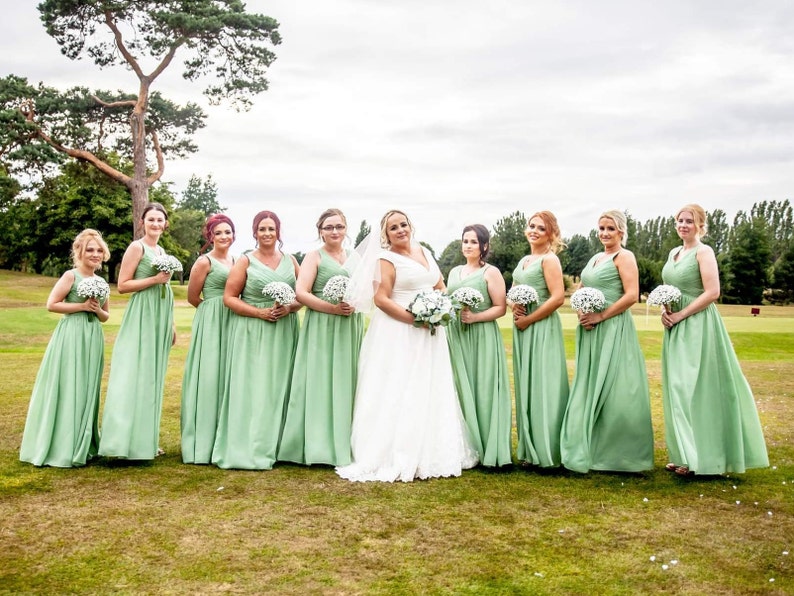 May include: A group of bridesmaids in sage green dresses stand in a line on a golf course. The bride is in the center wearing a white wedding dress. The bridesmaids are holding bouquets of white flowers.