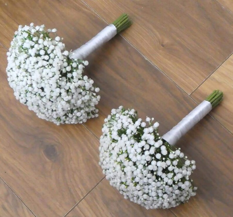 May include: Two white baby's breath bouquets tied with silver ribbon. The bouquets are arranged on a wooden surface.