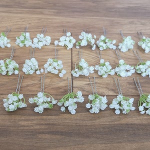 May include: Twenty-four white baby's breath flower hair pins with green stems. The pins are arranged in four rows of six on a wooden surface.