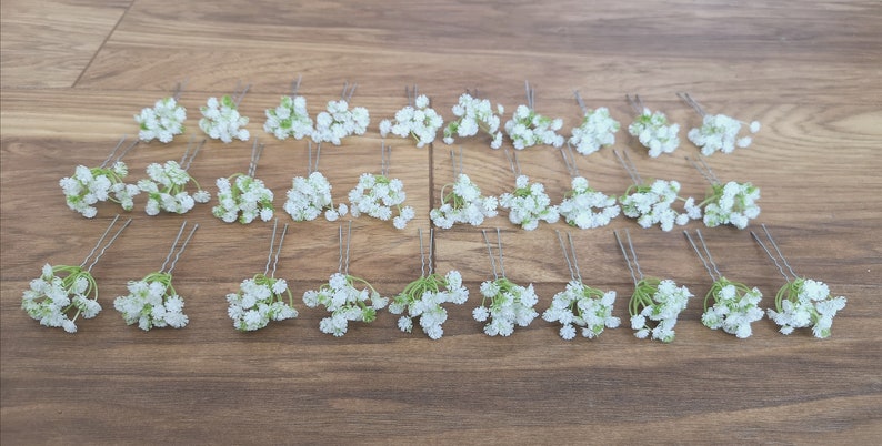 May include: Twenty-four white baby's breath flower hair pins with green stems. The pins are arranged in four rows of six on a wooden surface.
