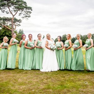 May include: A group of bridesmaids in sage green dresses stand in a line with a bride in a white wedding dress. They are all holding bouquets of white flowers and smiling at the camera.