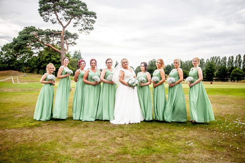 May include: A group of bridesmaids in sage green dresses stand in a line with a bride in a white wedding dress. They are all holding bouquets of white flowers and smiling at the camera.